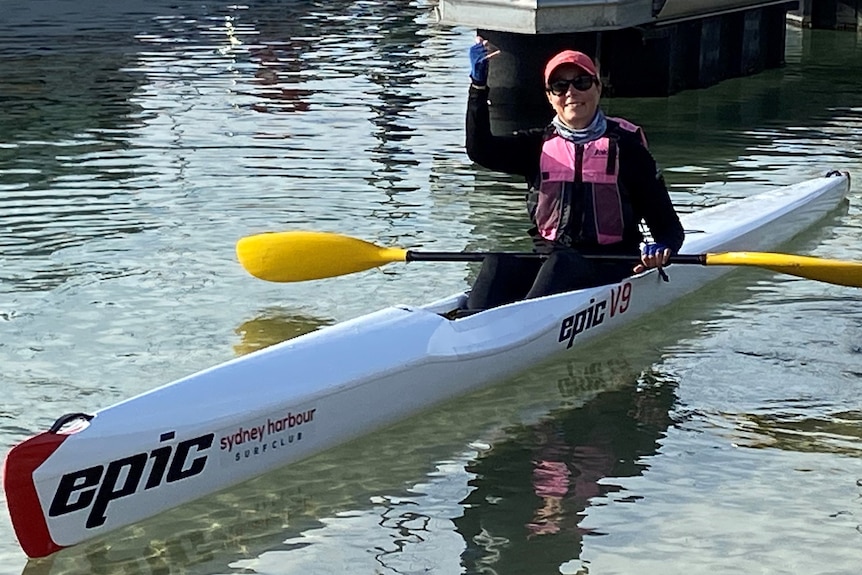 A woman with pink cap and pink life vest sits in a canoe on water waving and smiling widely.