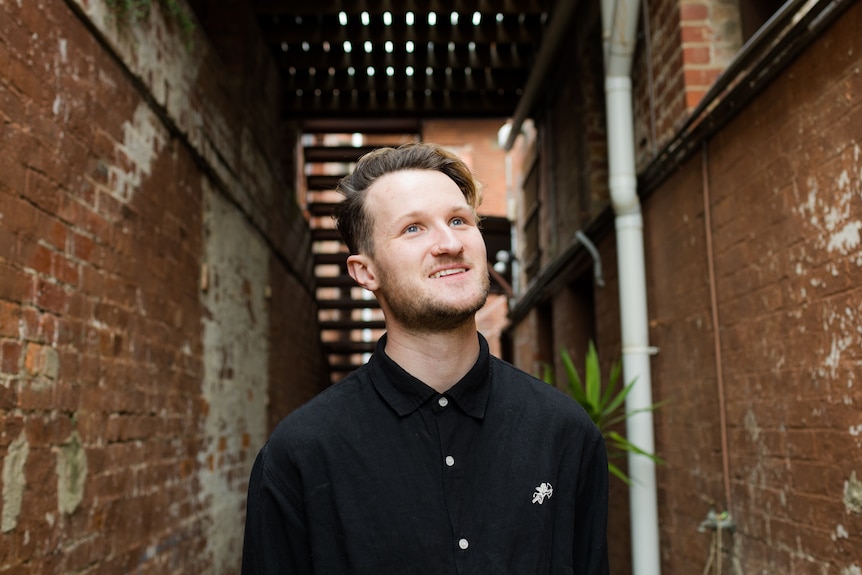 Blonde white man with light beard wears black button-up shirt and looks skyward in brick-lined alley.