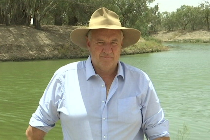A man in a collared shirt and akubra stands on a riverbank with the muddy river waters visible in the background.