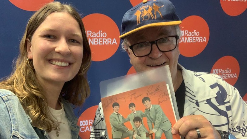 A woman and a man standing close to each other smiling and the man is holding a signed orange poster of the 4 Beatles