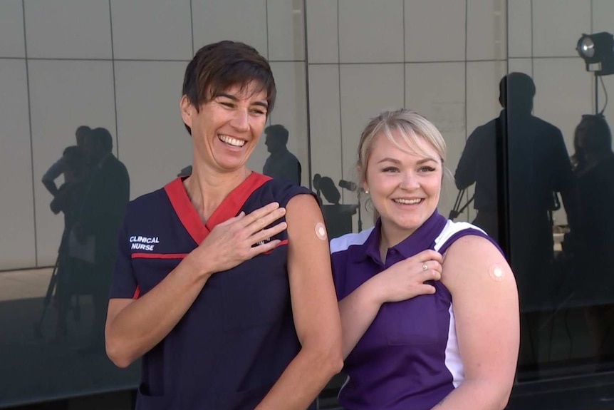 Two women in nursing uniform roll up sleeves to display small band aid over injection point after having COVID vaccine.
