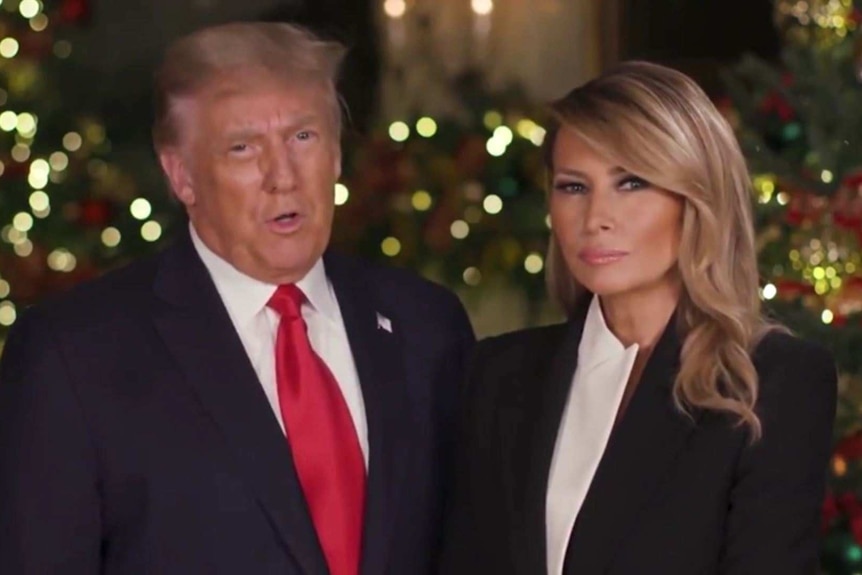 Donald Trump and Melania Trump in black suit jackets stand in front of Christmas Trees