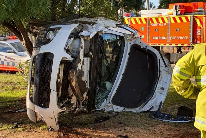 Image of white car on its side seriously damaged with emergency services in rear.