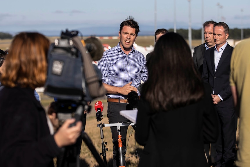 A man in a check blue shirt looks to the camera. He is surrounded by reporters and camera operators.