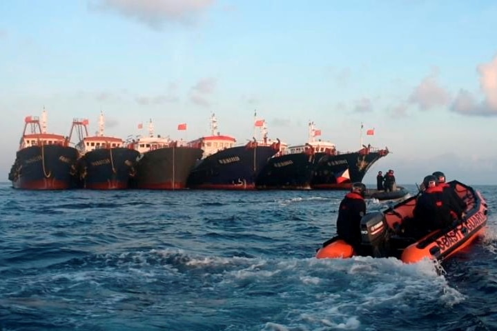 Three uniformed personnel ride in a small rubber boat over calm seas as they inspect a fleet of six large fishing vessels.