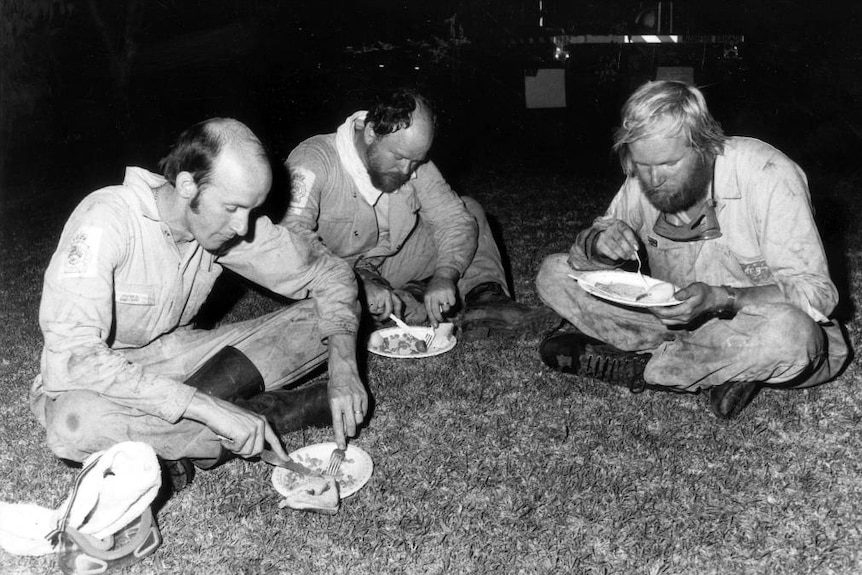 Firefighters sit on a lawn eating dinner