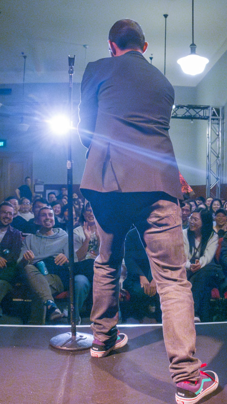 A stand-up comedian performs in front of a seated audience with his back toward the camera 
