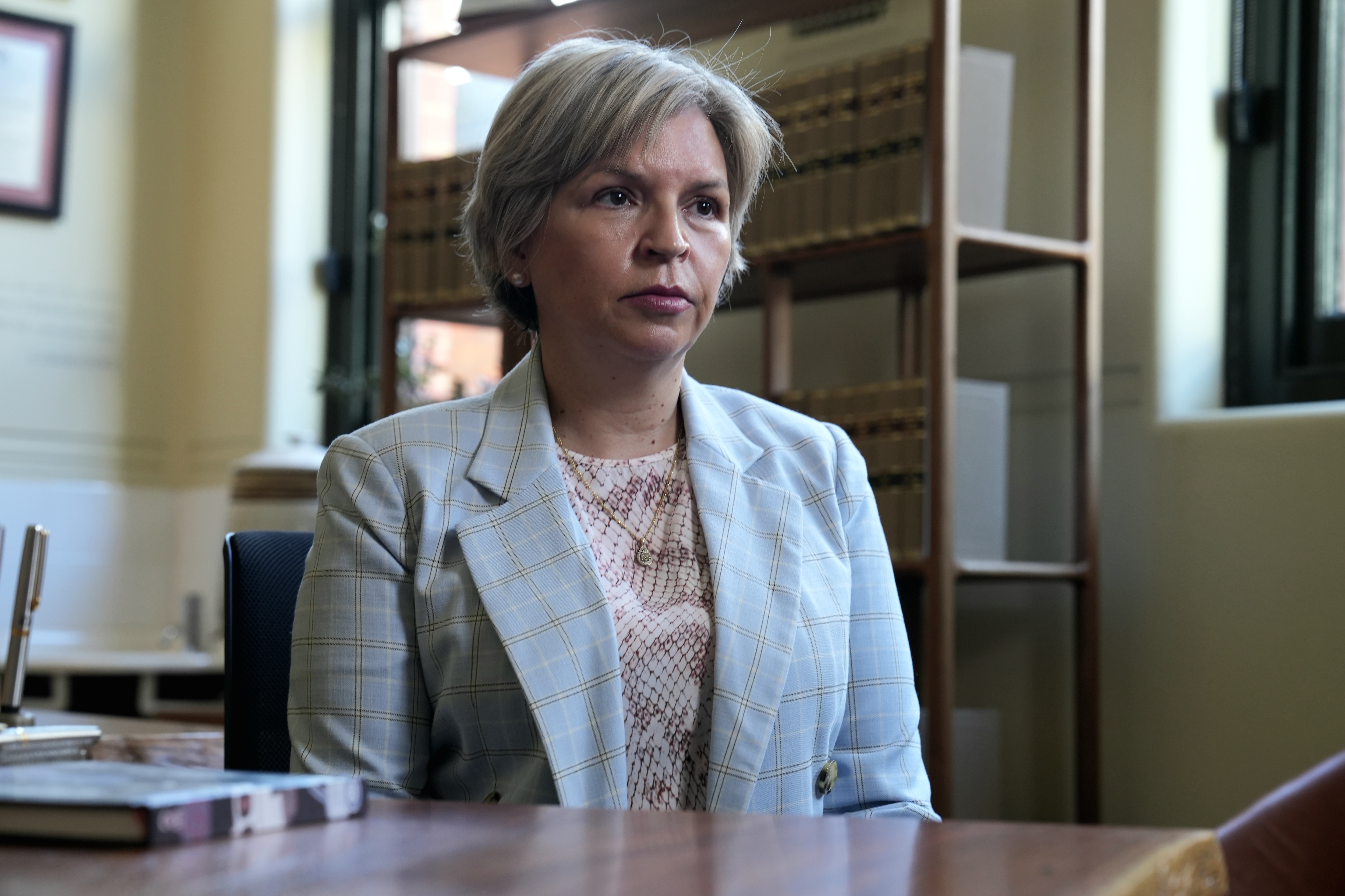 Una mujer con un traje beige está sentada frente a un escritorio de madera oscura.