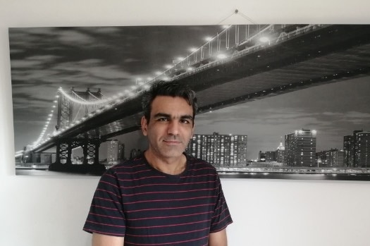 A grey haired man smiles, standing in front of a picture of Brisbane's Gateway Bridge.