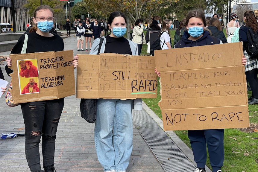 Three girls wearing face masks hold up signs protesting against rape and sexual assault