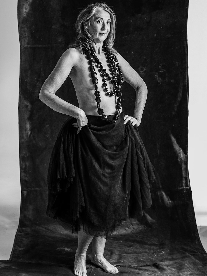 Black and white full length photo of woman with long hair, no top, necklace over breast, full length skirt, worn feet.