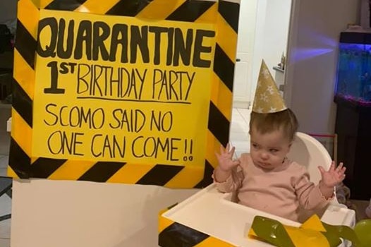 A baby sits in a high chair next to a yellow sign reading: Quarantine 1st birthday party, Scomo said no one can come!!