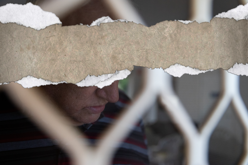 A photo of a man standing behind a flyscreen door with the top half of the photo ripped off, hiding his face.