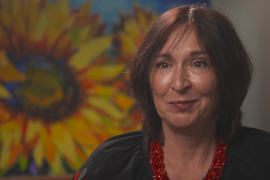 Woman with brown hair sitting behind a painting of sunflowers.