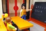 Lego figurines sitting at desks in a classroom, facing a figurine dressed as a teacher, which is pointing at a blackboard.