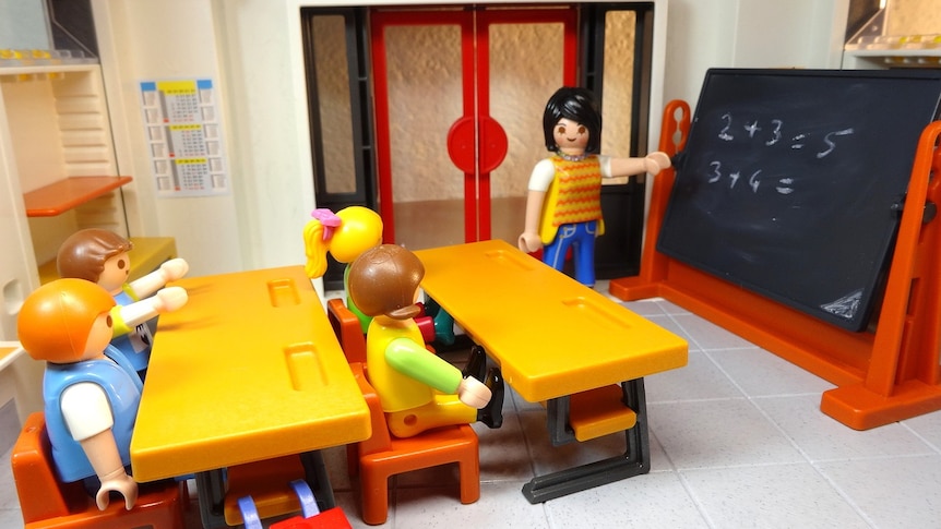 Lego figurines sitting at desks in a classroom, facing a figurine dressed as a teacher, which is pointing at a blackboard.