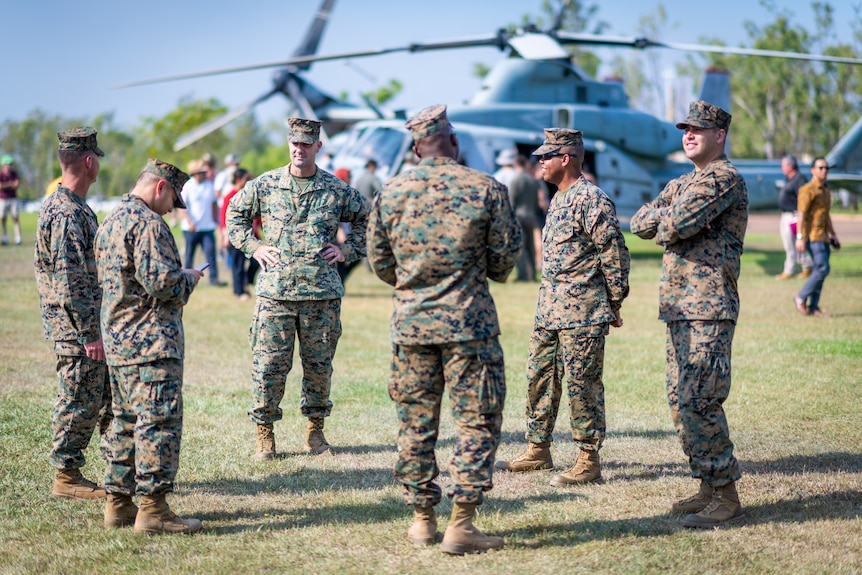 Male marines in uniform stand in a circle talking. A plane is in the background.