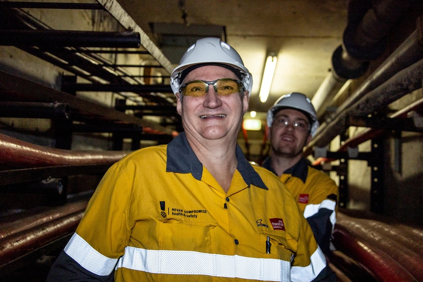 Mal Harrison smiles, dressed in high-vis and a hardhat, in a dark tunnel lined by pipes.