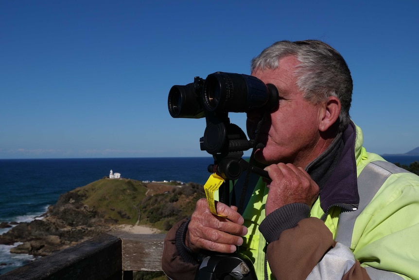 A man with grey hair, wearing bright yellow jacket, looking through binoculars with the ocean in the background.