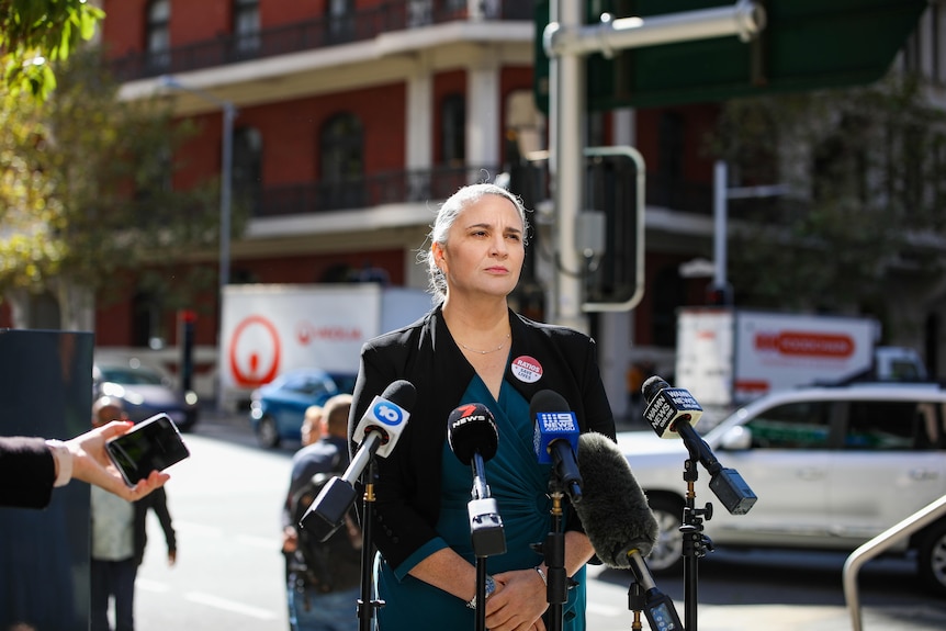 A woman wearing a dark blazer looks serious in front of microphones. 