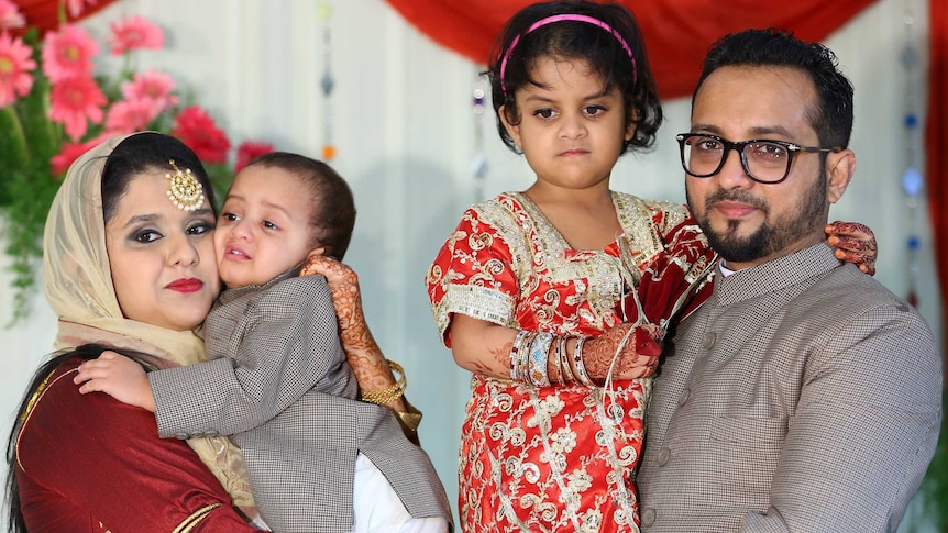 Siraj Patel stands holding his daughter next to his wife who is holding their son