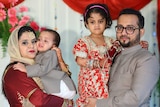 Siraj Patel stands holding his daughter next to his wife who is holding their son
