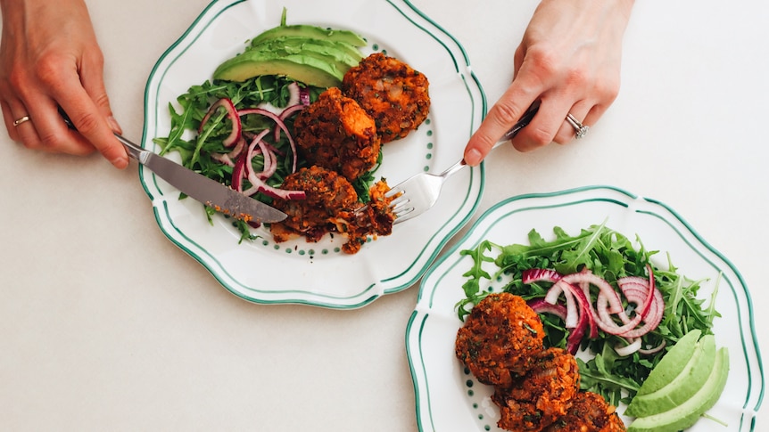 Two plates piled with orange coloured veggie patties and salad, a pair of hands hovers over holding knife and fork