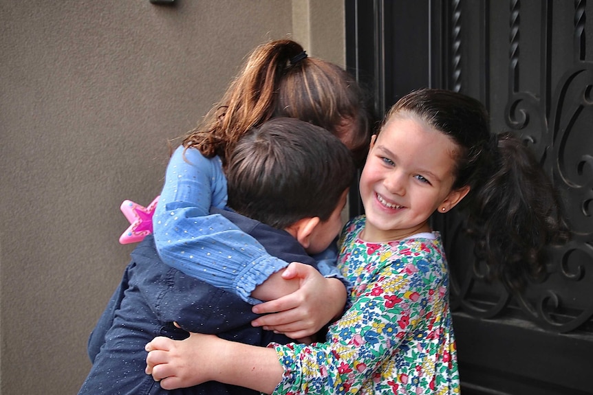 Three young children, two girls and a boy embrace outside a door.
