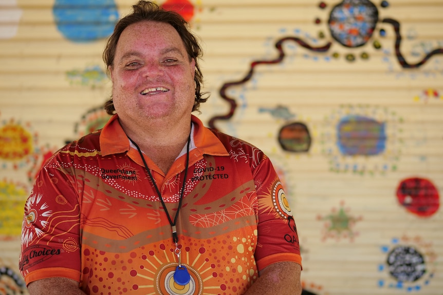 Terrence standing in front of Aboriginal artwork, smiling, wearing red and orange colourful shirt.