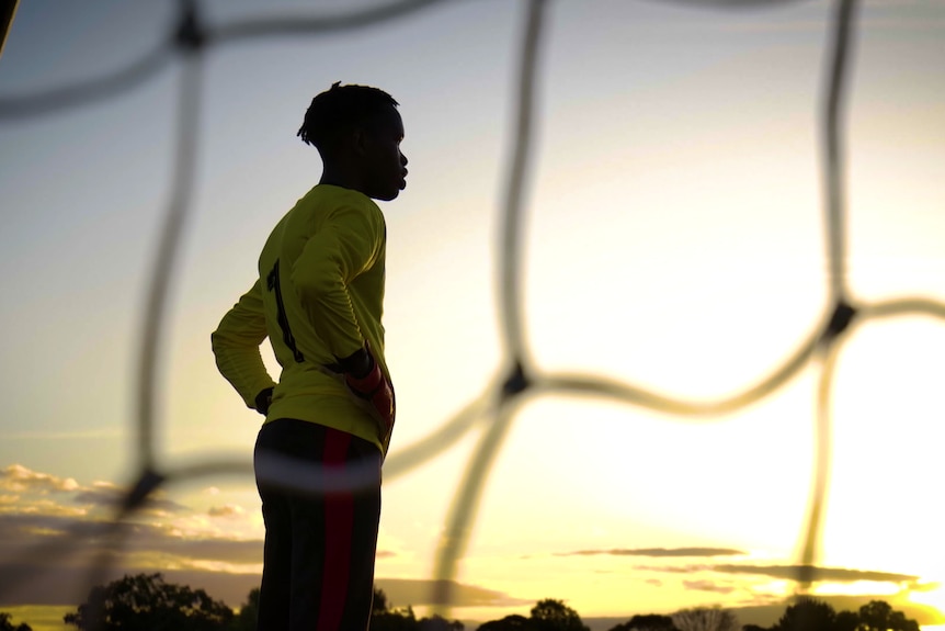 A silhouete of a young black person in front of a soccer goal. He is looking out to the horizon, where the sun is shining