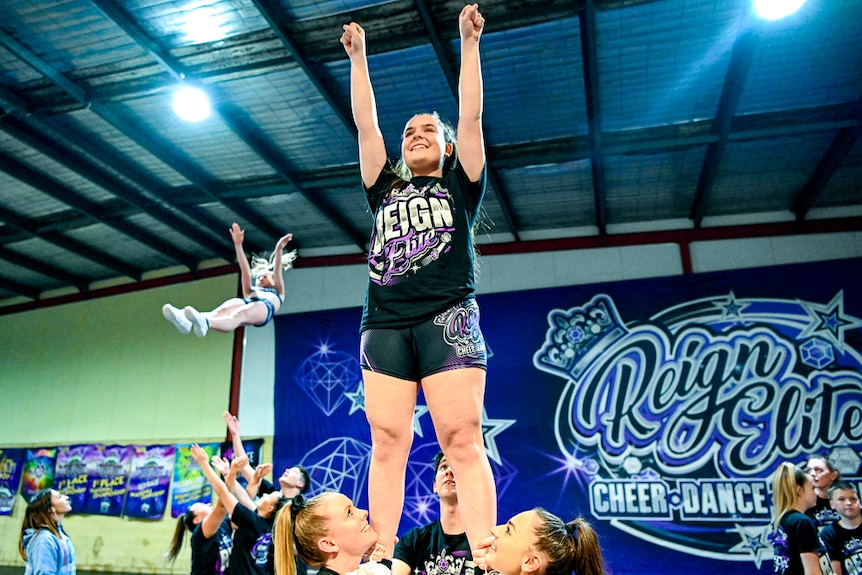 A teenage girl is held in the air by three athletes while she poses with both arms straight up.