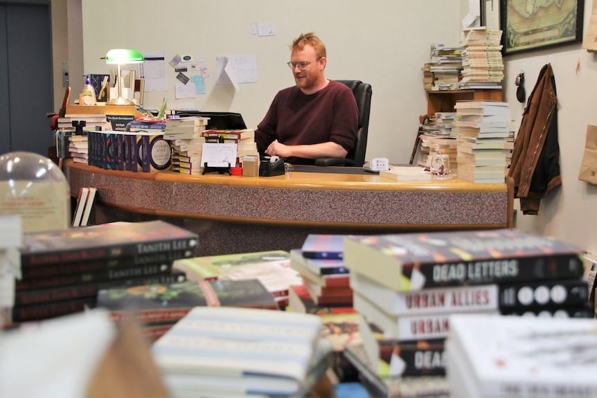Toby Wools-Cobb works away at his desk, surrounded by towering piles of books.