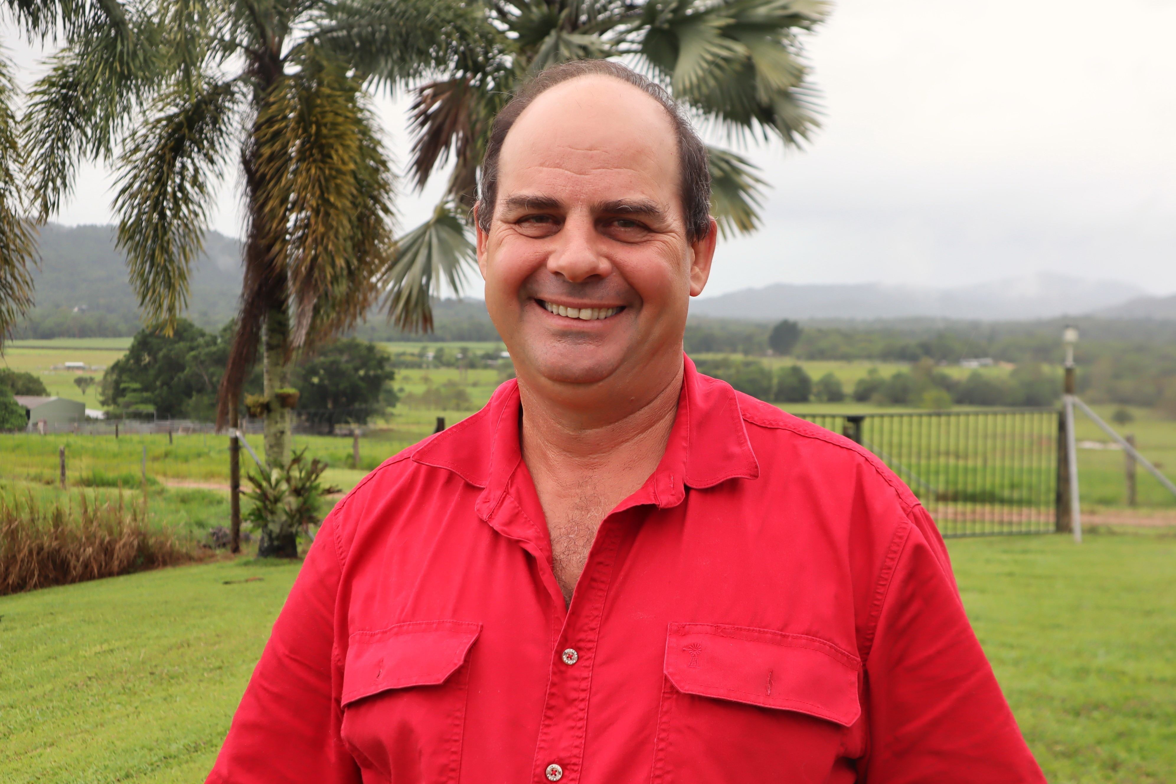 A man with balding hear wears a red shirt and smiles standing in a green paddock.