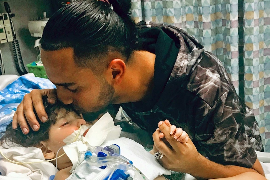 A man kissing his dying two year old son on the forehead in a hospital.