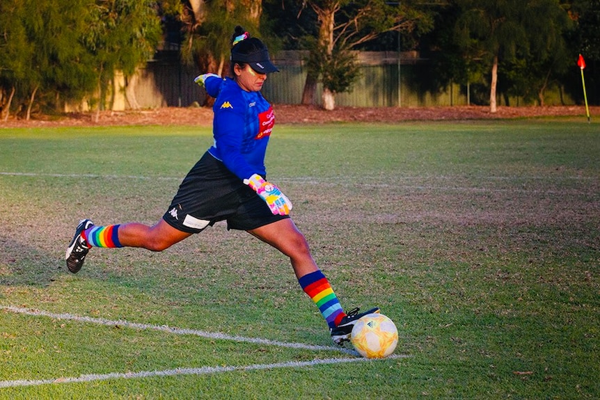 Aish Ravi kicks a soccer ball in rainbow coloured socks a blue jersey and black shorts.