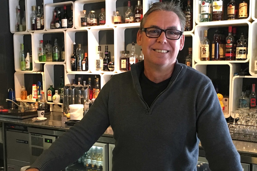 Pure Bar owner Michael Cutler in sweater and glasses, with shelves of alcohol behind him.