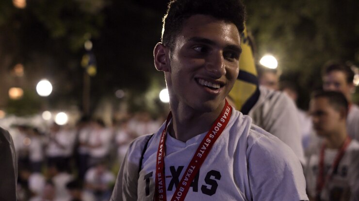 A male teen smiles to his left side during night time rally or gathering in park with large crowd of boys all in white t-shirts.