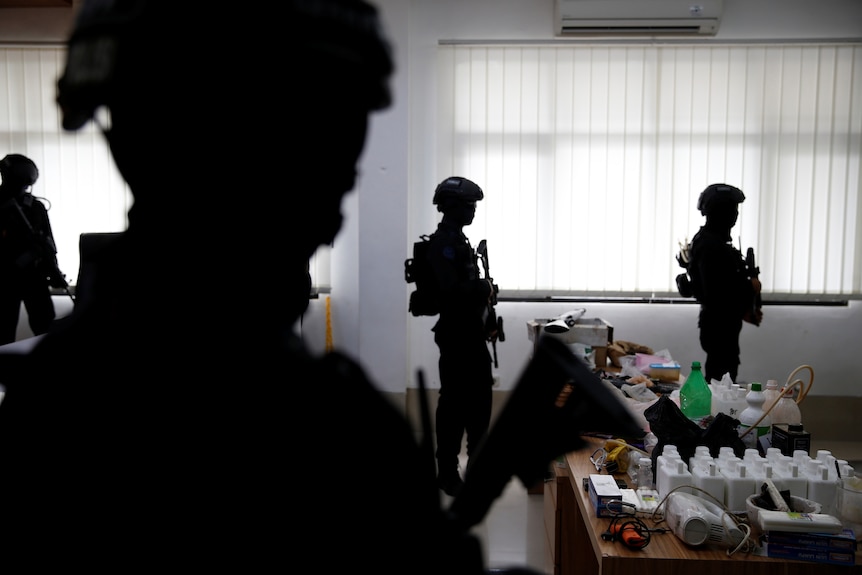 Silhouettes of policemen in helmets and guns stand near a table with evidence.