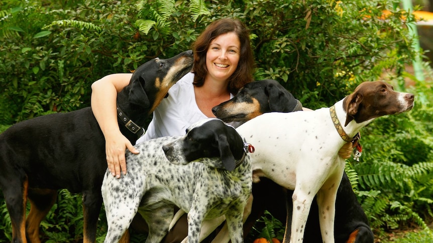 A smiling woman with shoulder length brown hair kneeling in front of a bush in between four dogs.