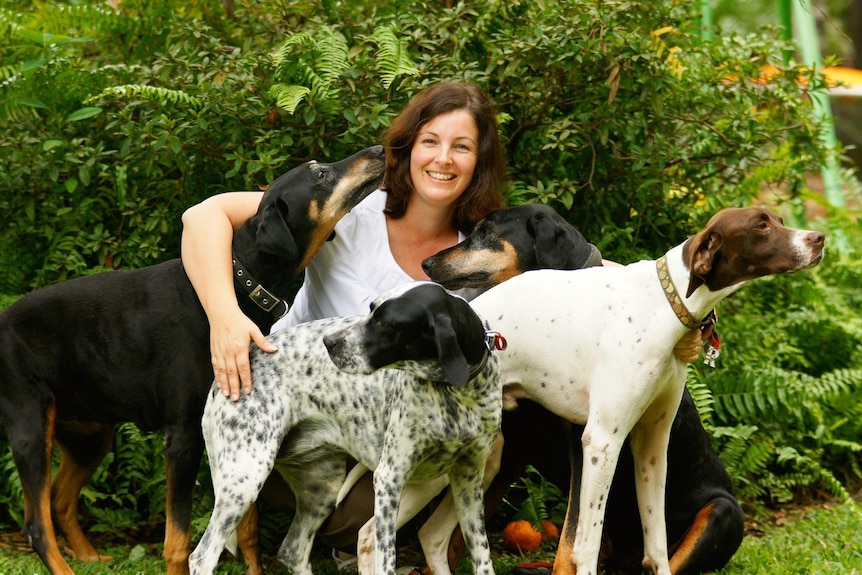 A smiling woman with shoulder length brown hair kneeling in front of a bush in between four dogs.