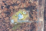 An aerial view of a house and outbuildings on a green patch, surrounded by burnt bushland.