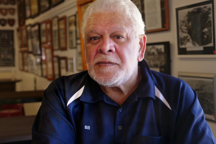 An older, bearded man with white hair sits in a club in front of memorabilia.