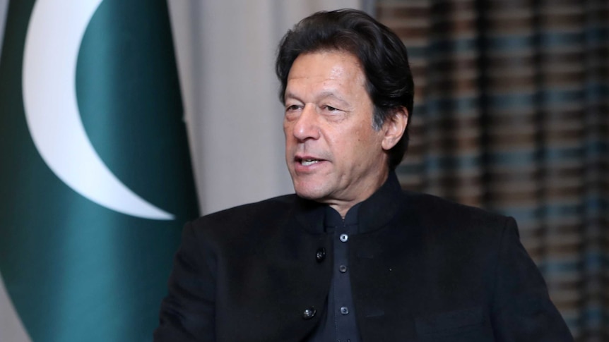 imran khan sits wearing formal shirt and blazer facing side on in front of pakistan flag and curtain in background