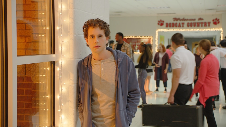 A young man looks distressed, leaning against a school hallway wall and looking into the camera lens