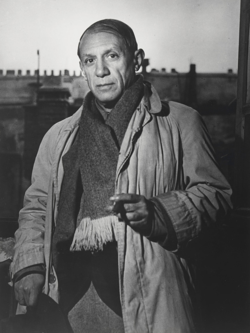 Black and white photograph of Picasso in his late 50s, balding, wearing heavy overcoat and scarf, standing in studio.