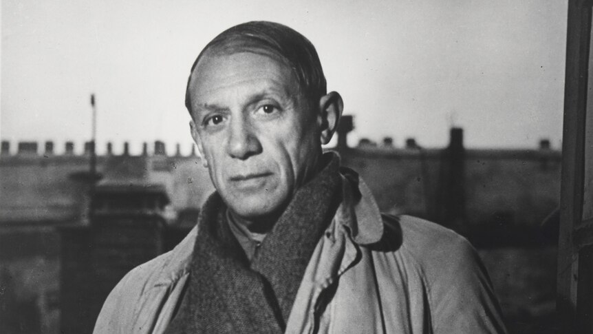 Black and white photograph of Picasso in his late 50s, balding, wearing heavy overcoat and scarf, standing in studio.