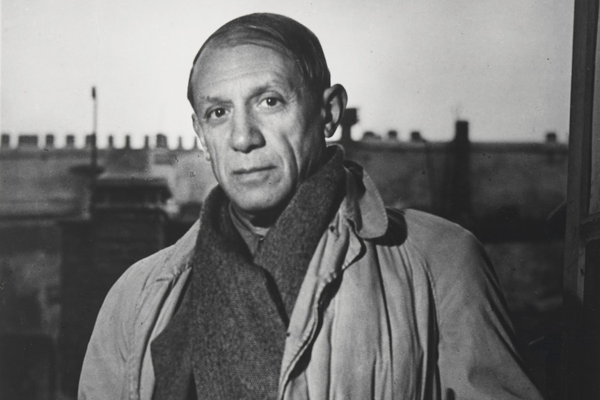 Black and white photograph of Picasso in his late 50s, balding, wearing heavy overcoat and scarf, standing in studio.