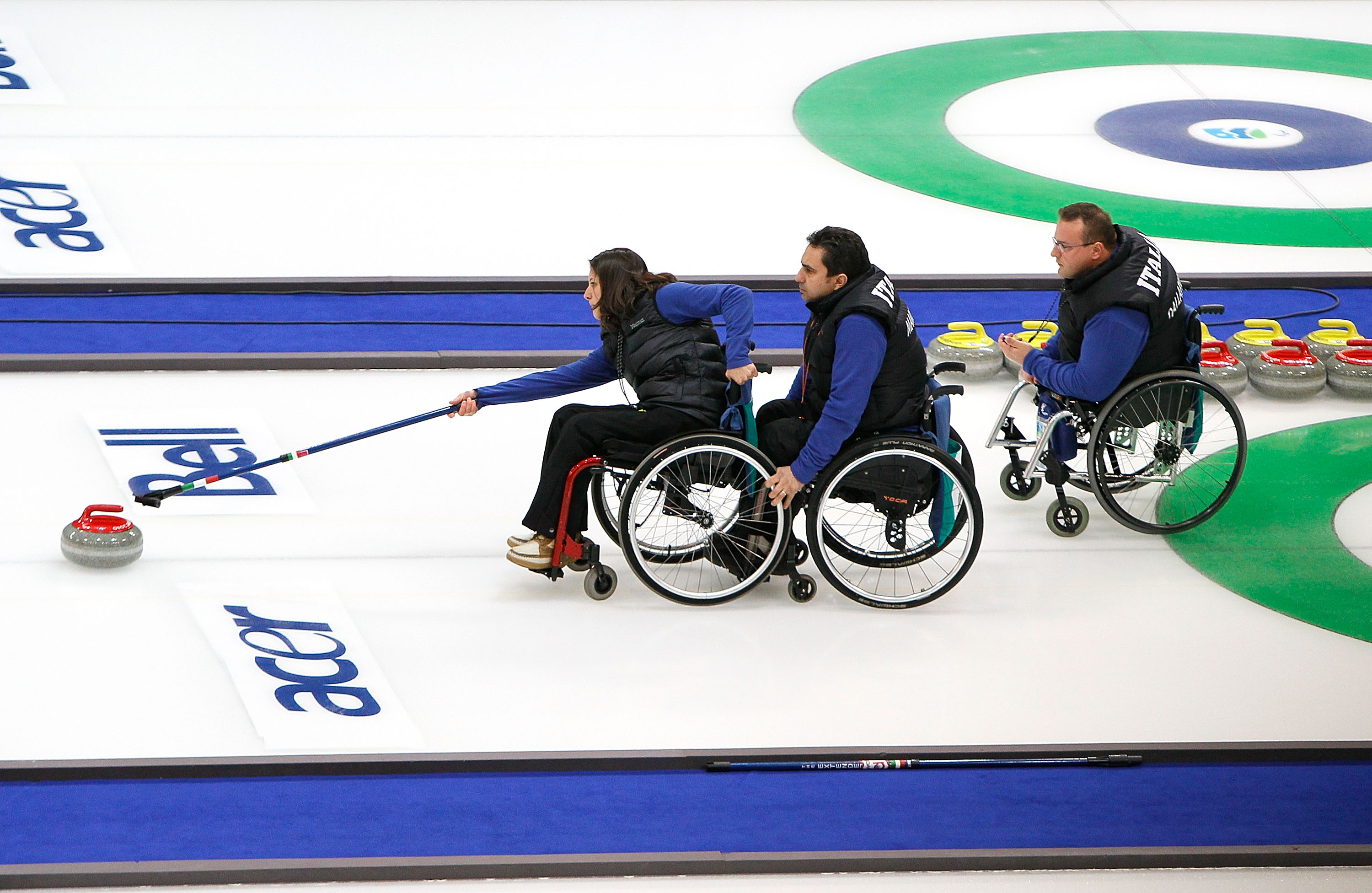 Três atletas que usam cadeiras de rodas estão concentrados enquanto empurram uma pedra no gelo