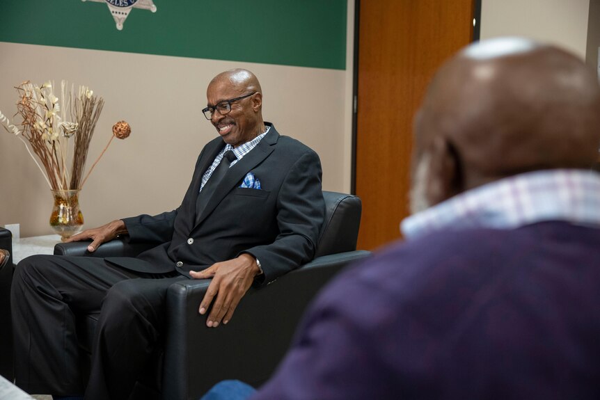 A bespectacled black man in a suit sits grinning in an arm chair