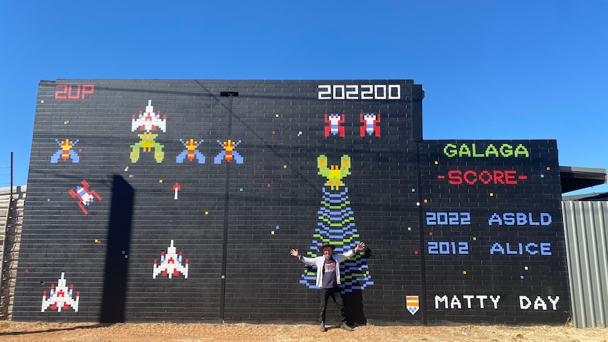 A man stands with his hands in the air in front of a mural featuring 80s style arcade game aliens.